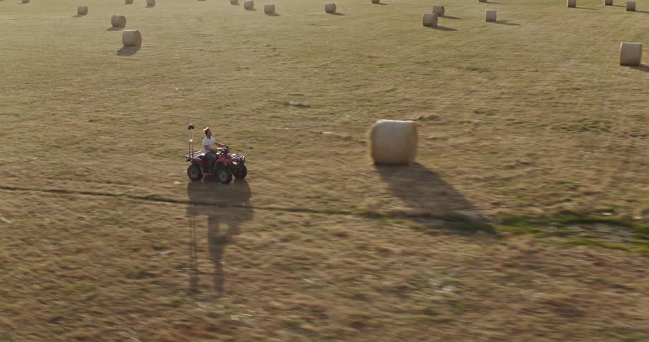 A man rides an all terrain four wheeler through a vast field containing rolls of hay during the day