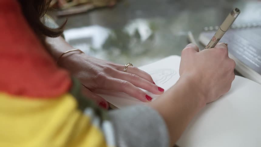 A woman sits at a table and writes in a blank notebook with a pen