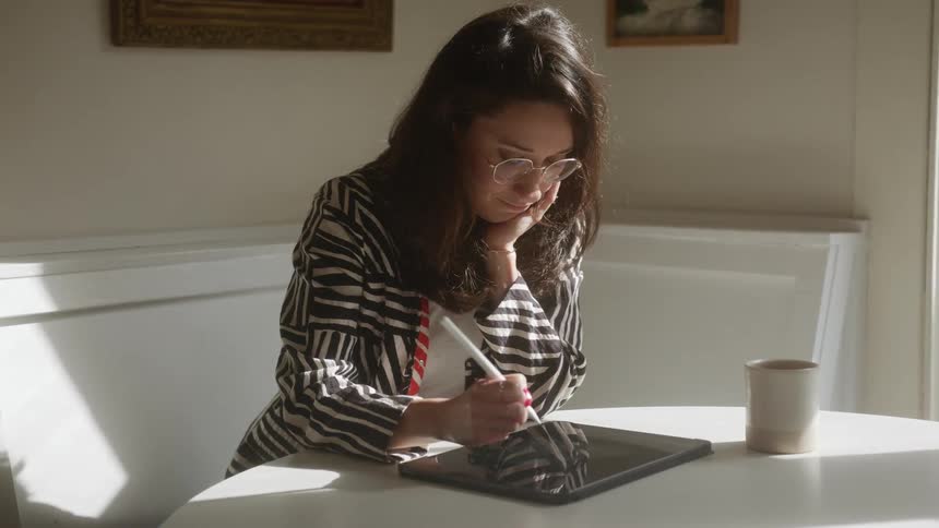 A woman sits at a table and concentrates on using a stylus along with her tablet computer