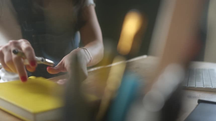 A woman sits at a table and places pens on top of a notebook and reaches for a laptop