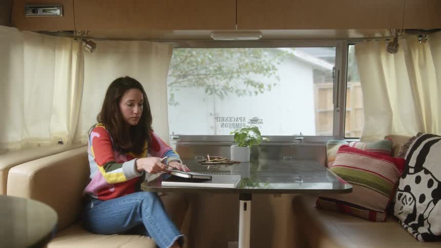 A young woman sits down with a book at a table inside a camper