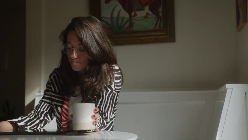 A woman sits indoors at a table and takes a drink from a large coffee mug