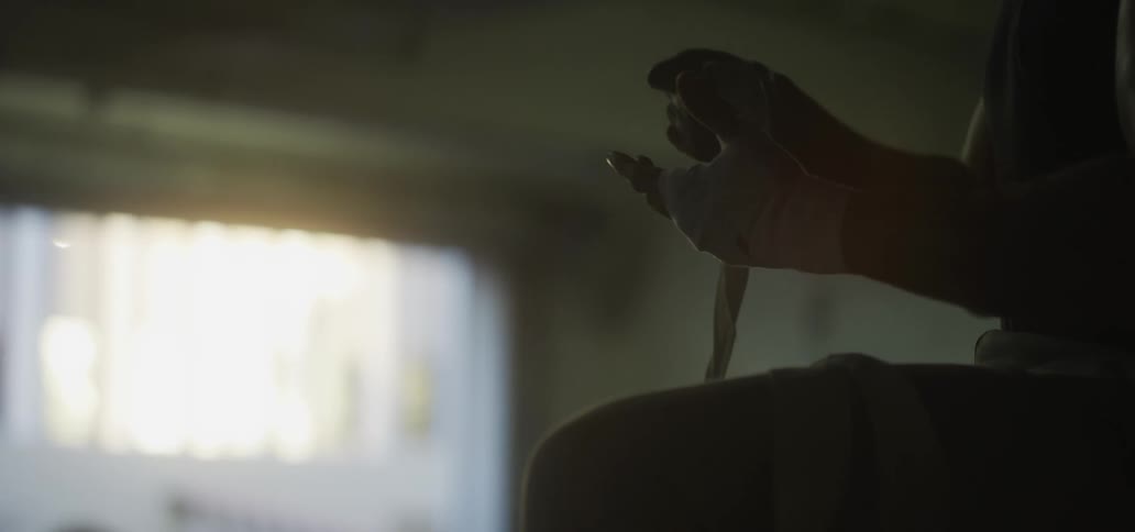A man sitting in a dimly lit room slowly unwinds the handwraps on his hand