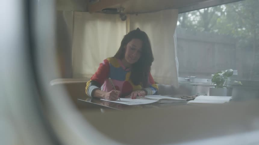 A woman uses a marker to draw in a notebook while sitting at a table in an rv trailer