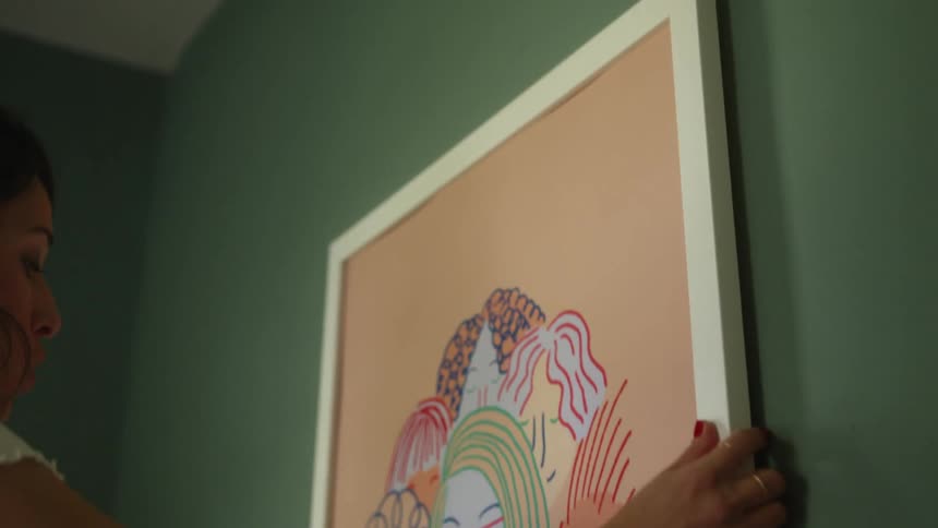 A woman lifts a painting off of a leather sofa and hangs in on the wall above the sofa
