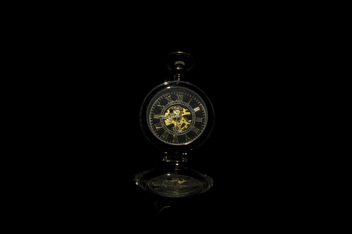 A gold clock with a stand is in darkness with the clock hands continually moving in a clockwise rotation