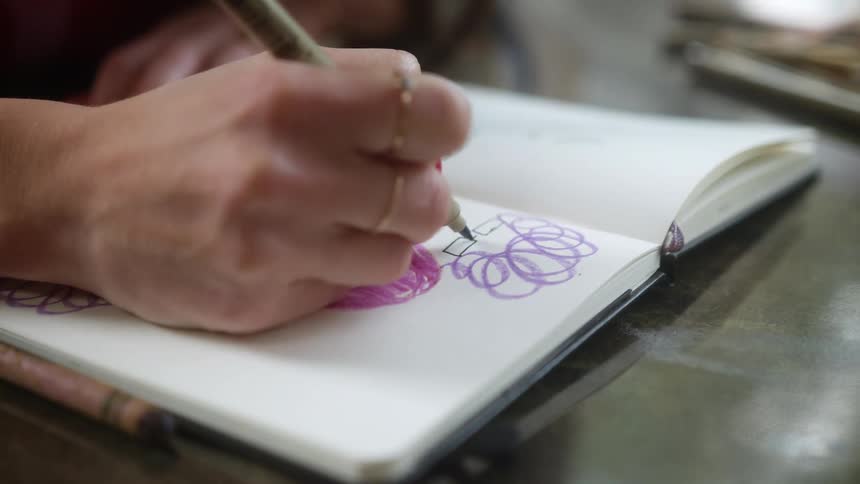 A woman doodles with a pen in a notebook sitting on a table