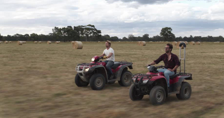 Two men drive quads across a farm field with bales of hay in it