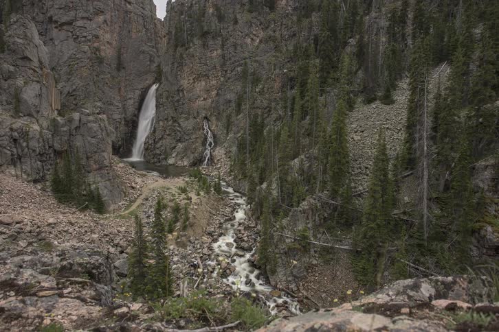 Time lapse of a side of a mountain with a small waterfall pouring down into a river while people walk about down below