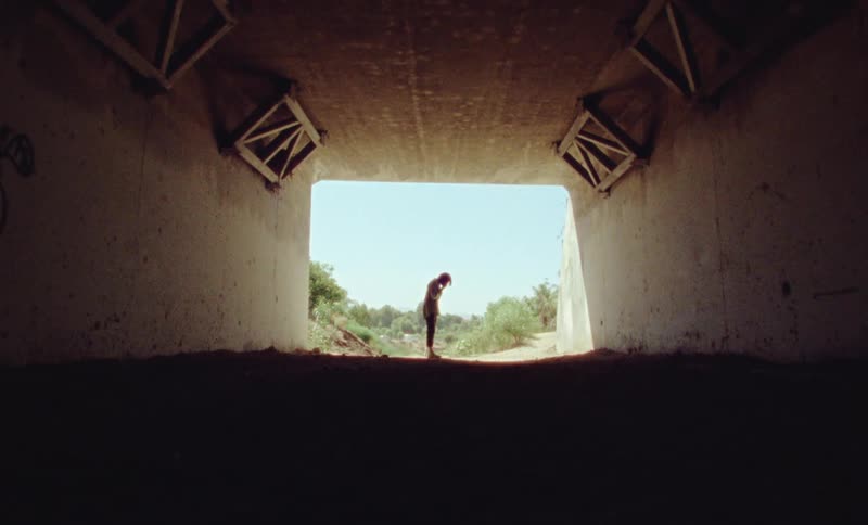 Woman talks on phone near mouth of dirt floored enclosure with scrub covered slope and clear blue sky behind her