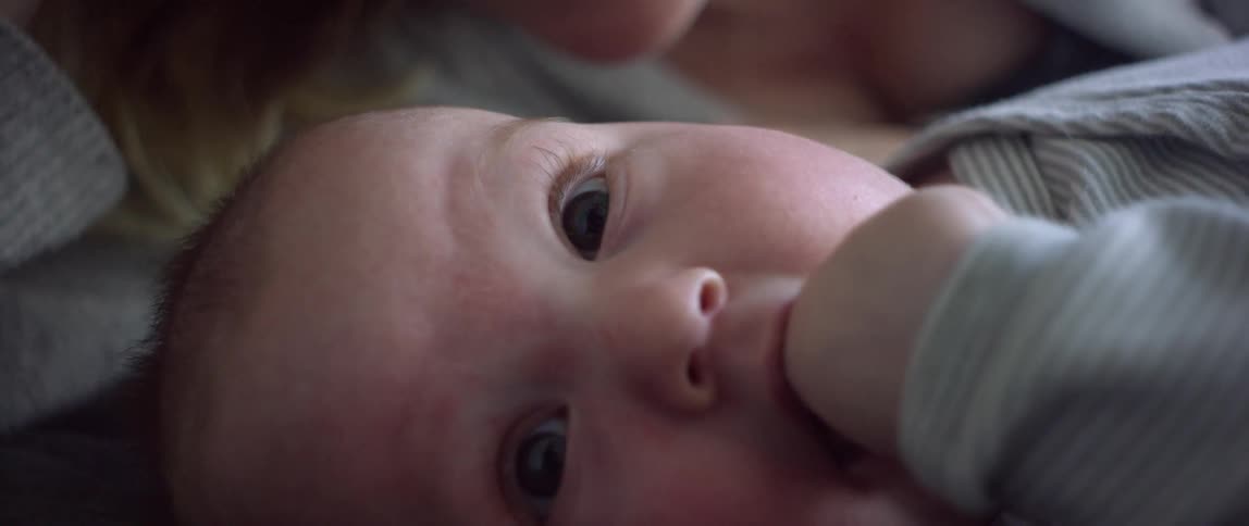 A baby lying down with his fist in his mouth in front of his mother