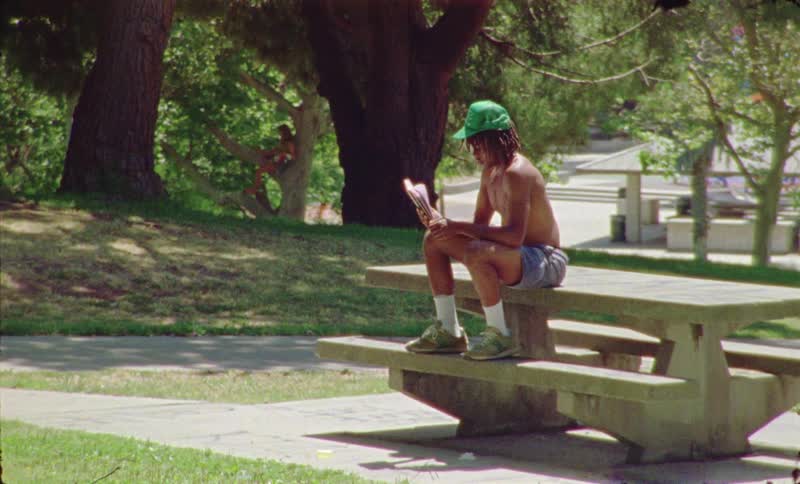 A bare chested man wearing shots and a green hat sits on a table in a park oudoors while reading a book