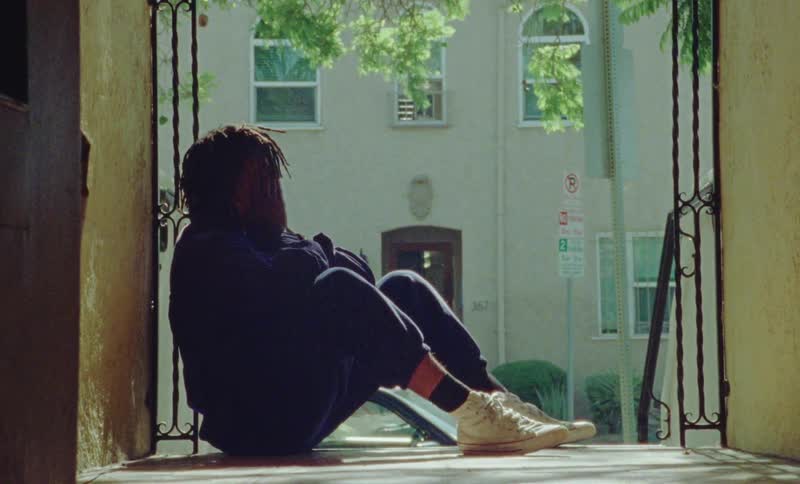 A person sits while covering their face with their hands on the top of a staircase in front of a building in a neighborhood during the day