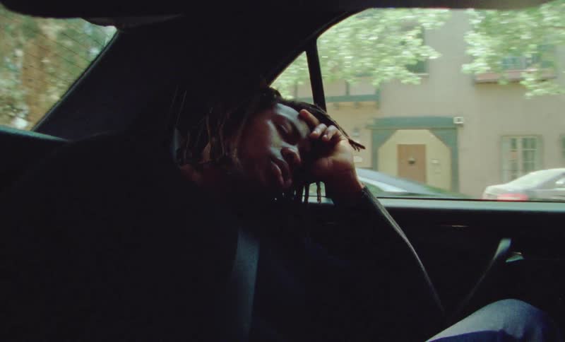A backseat passenger rests his head against the window of a car as it passes through a neighborhood filled with apartment buildings