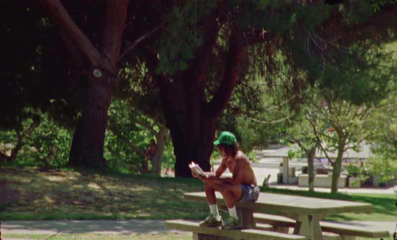 A bare chested man wearing shorts and a green hat sits on a picnic table in a park surrounded by trees holding a book during the day