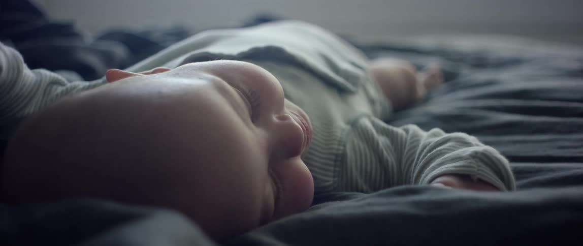 A baby lies on its back on a bed covered with a blue blanket moving hands and legs while smiling