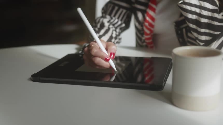 A woman with red nails writes on a tablet while sitting at a white table