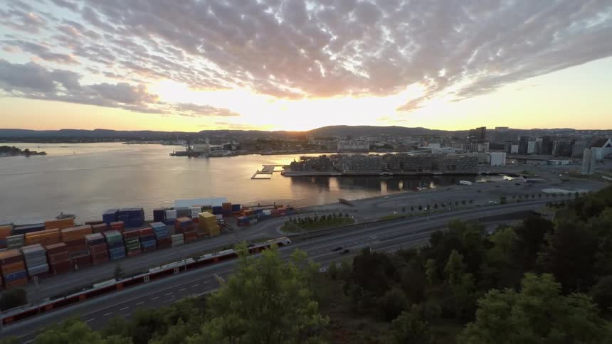 A view moving over a hill across a highway with a train and toward a busy port