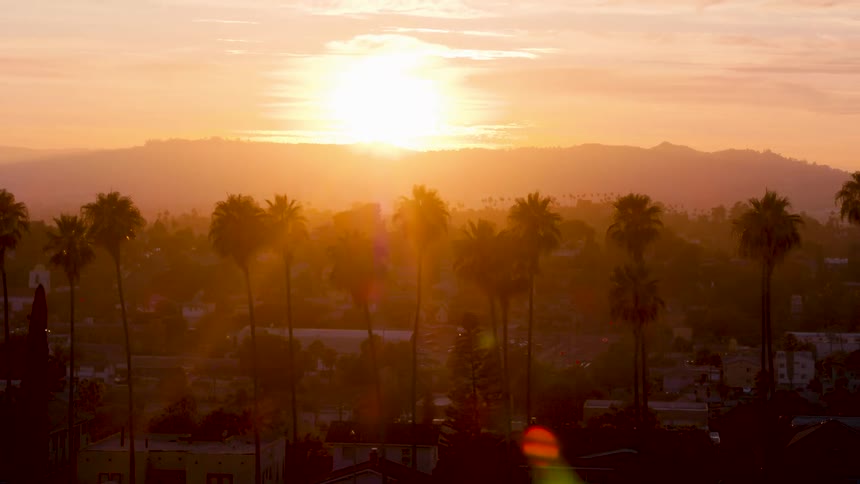 Huge sun setting in orange sky over mountains with palm trees in the forefront