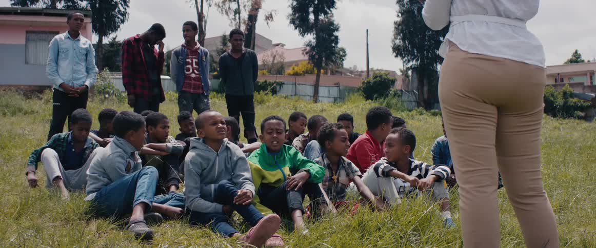 A woman stands in front of a group of seated children and standing teenagers on a lawn
