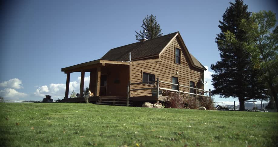 A wooden house sits on a field of grass with a couple of trees against a blue sky in sunny weather