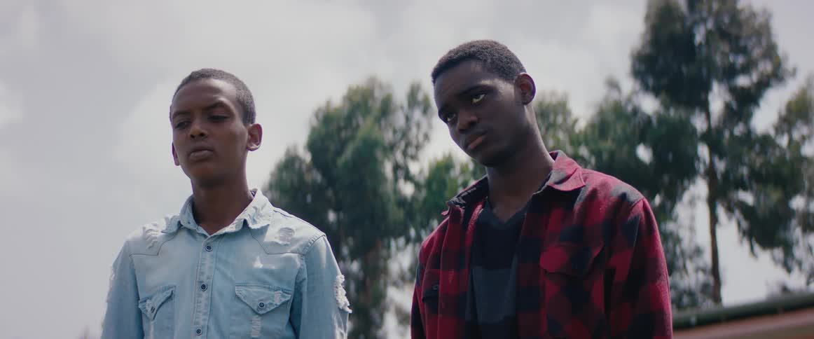 Two male teenagers stand outdoors with trees behind them