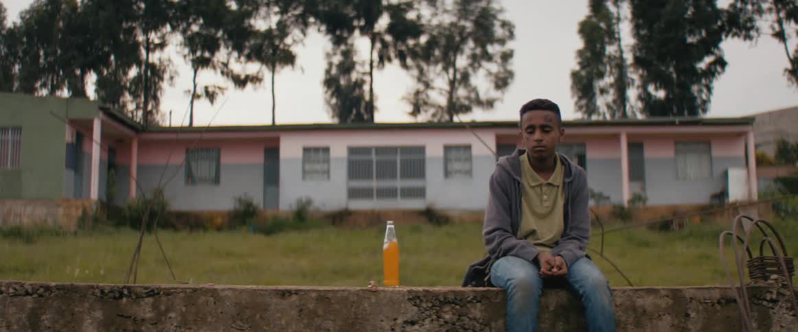 Black boy sitting on ledge reaching for bottle cap near orange soda outside