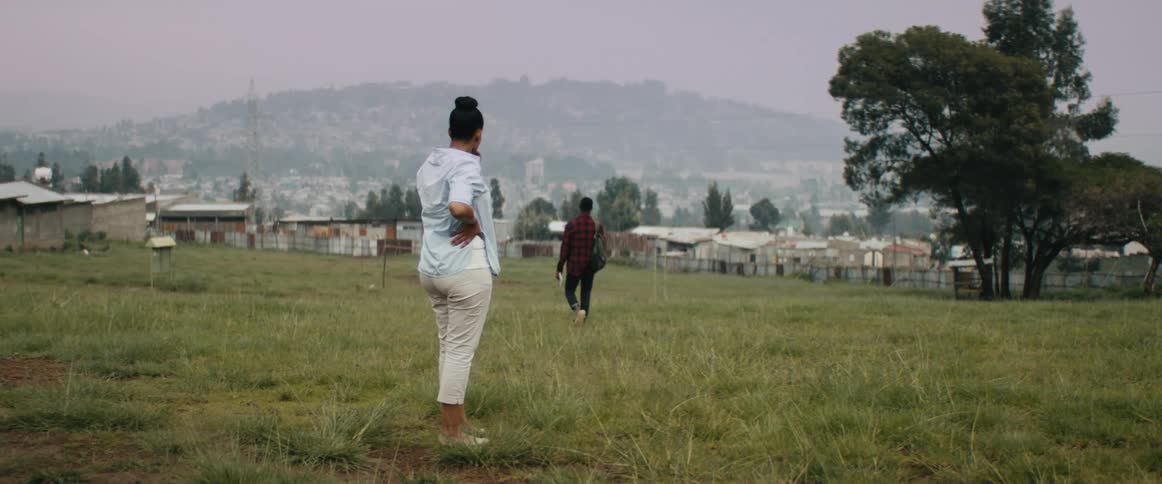 A woman stands outside in a park watching another person walks toward a neighborhood