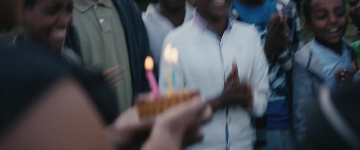 A group of boys standing in a circle in a field clap and sing while an older woman holds a cupcake with lit candles on top