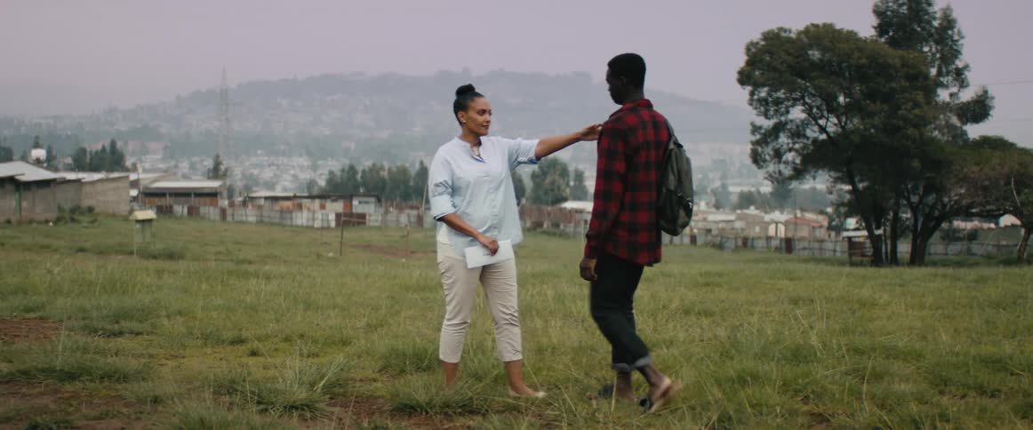 A young man walks up to his mother in a field near a large neighborhood and gives her a hug