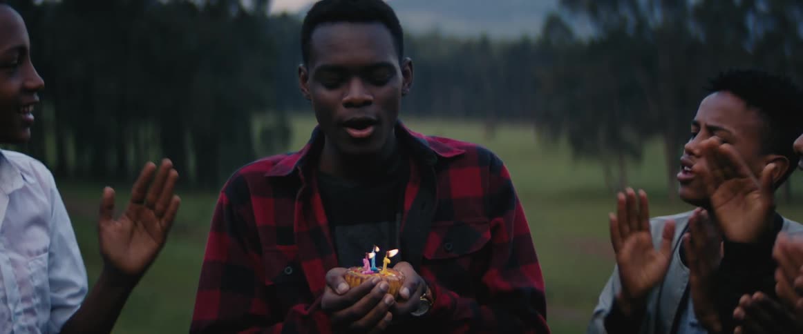 A man blows out candles on a cupcake he holds in his hands while boys clap and celebrate next to him outside