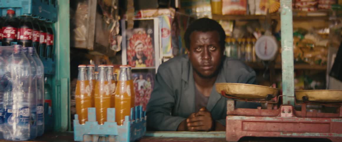 A shop keeper sits at a counter with orange soda and water bottles on a counter
