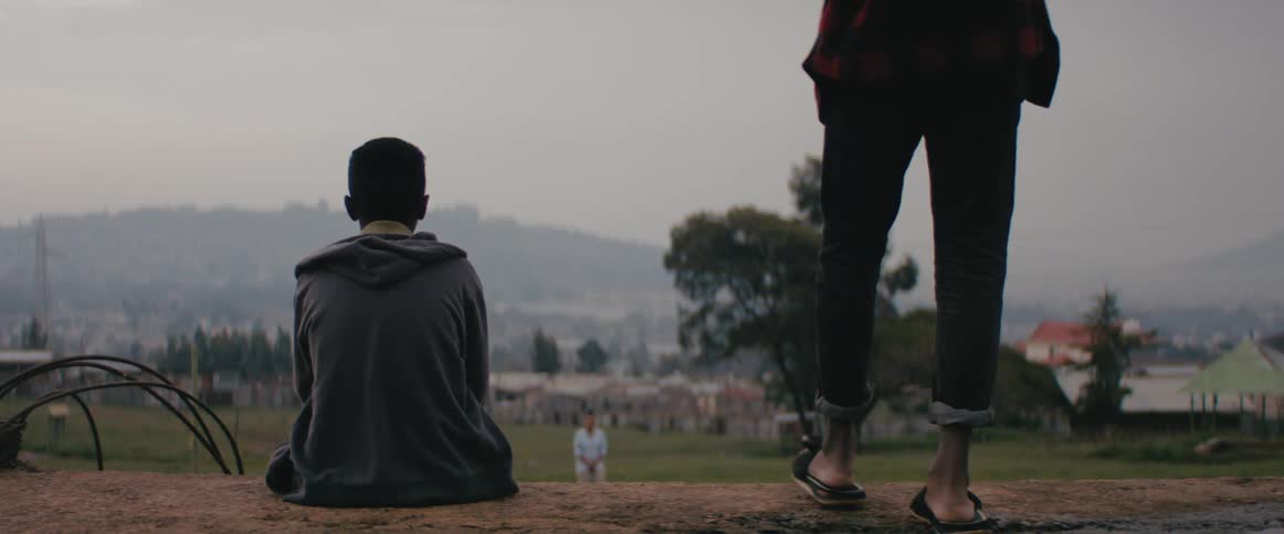 A boy sits down outside in grass while a man with a backpack walks over to him and joins him