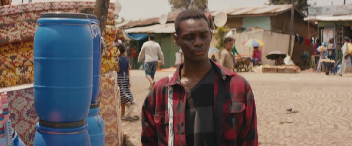 A man in a plaid shirt is among many people walking down the street in a developing city as vehicles pass by
