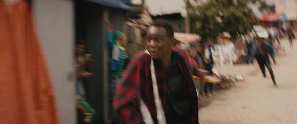A man wearing a red plaid shirt is being chased by another man on a street as people watch