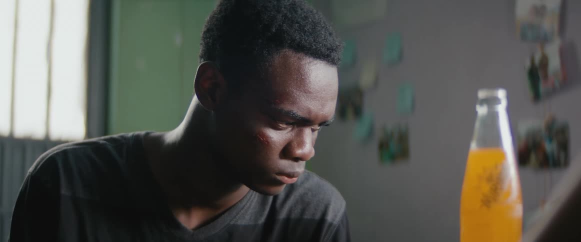 A young black man sits indoors and focuses intensely with a bottle of orange soda nearby