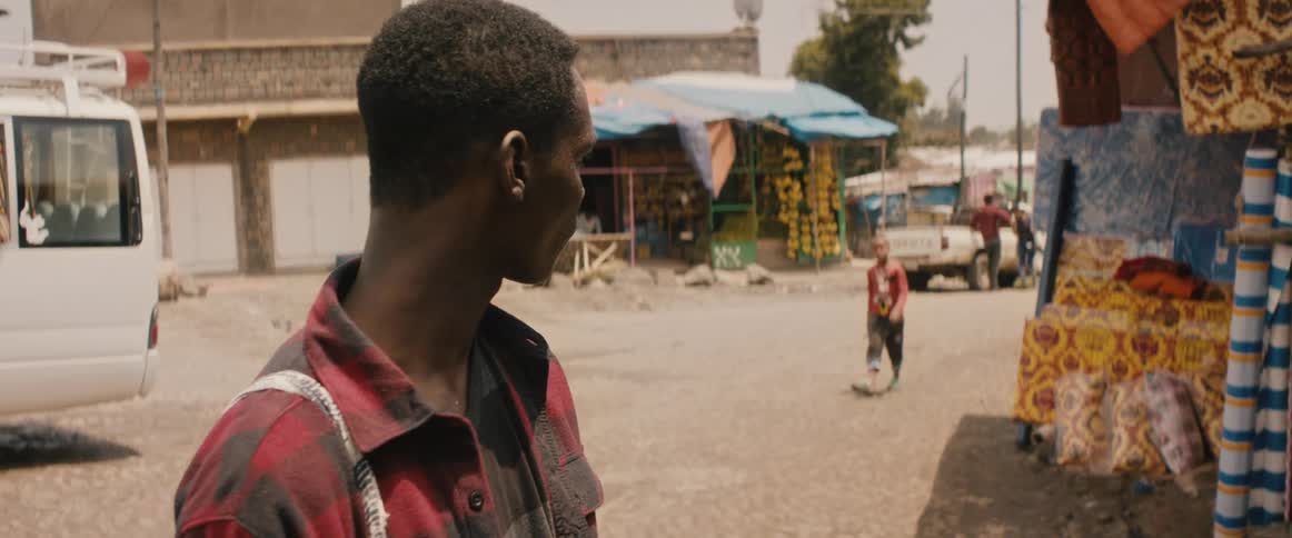 A teenage boy looks around nervously as he stands outside an outdoor shop in a village