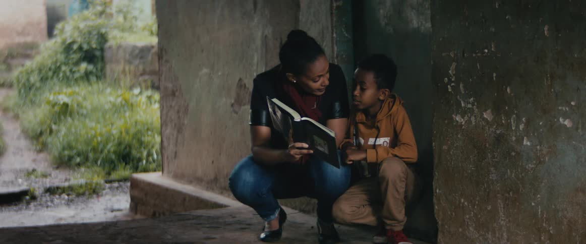 Mother crouches near sitting boy as they read from books near building with overgrown yard