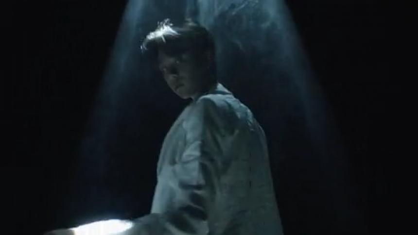 A asian female in a long white overcoat stands in a dark room under beams of light