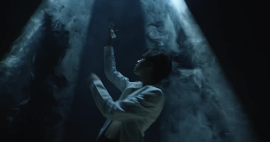An asian man wearing a white jacket stands in darkness with an above lightilluminating him