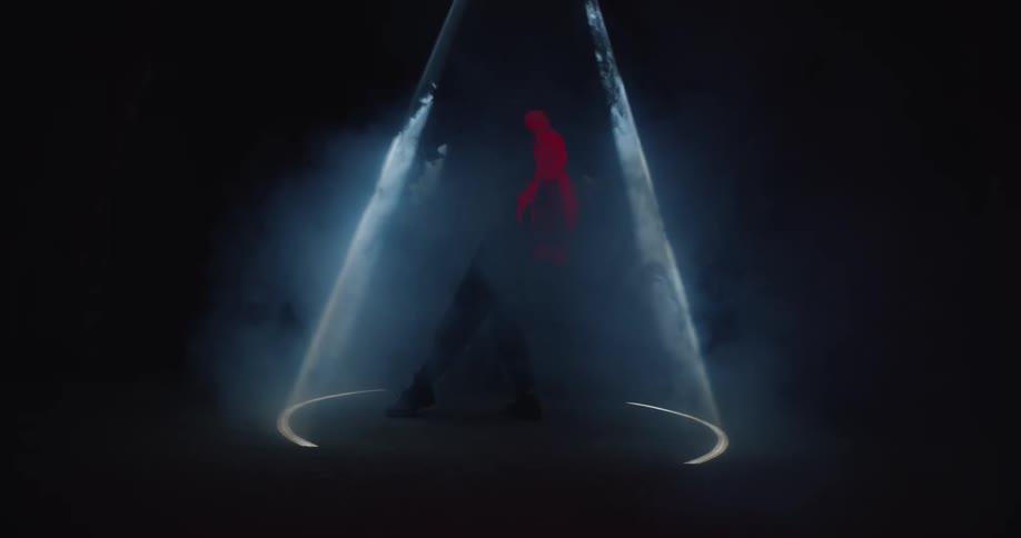 A circular light shines above a person wearing a red jacket who dances underneath it