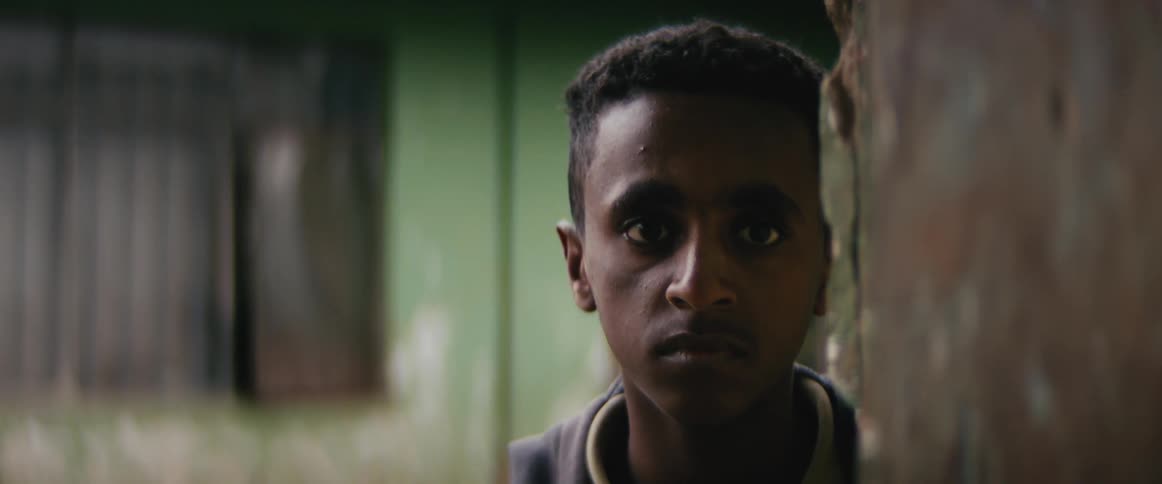 A young man standing outdoors in a village stares into the camera
