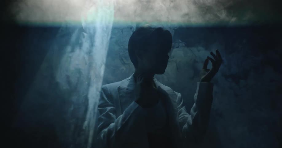 A model in a white blazer and sports bra looks at her hands and moves them around as a light from above flashes over her
