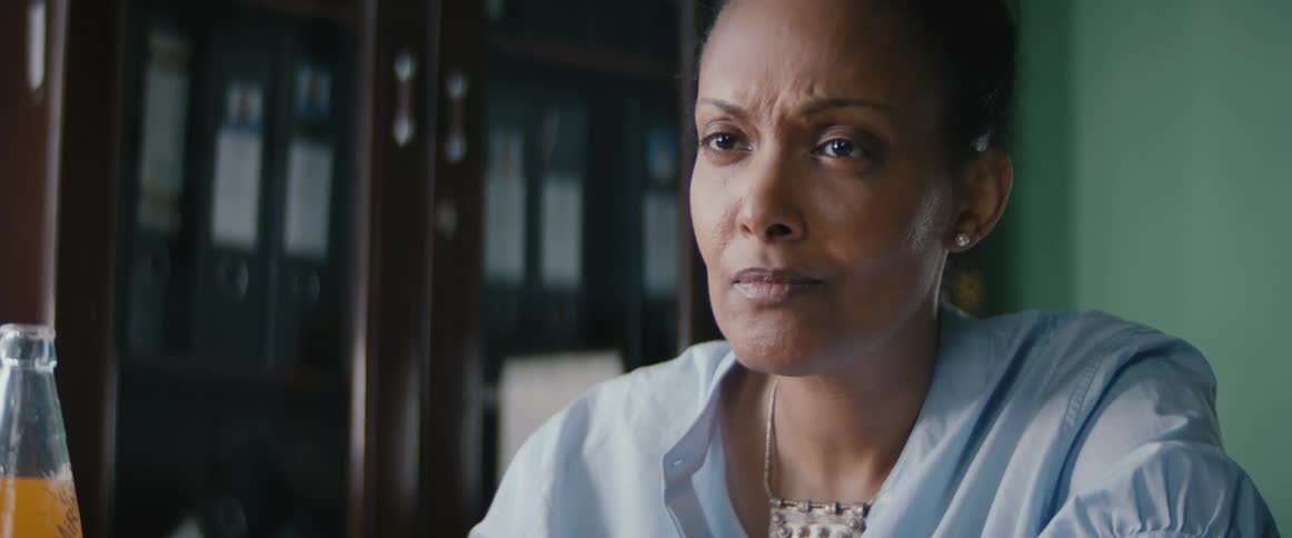 A woman sits at a table with an orange soda bottle shaking her head with a stern facial expression