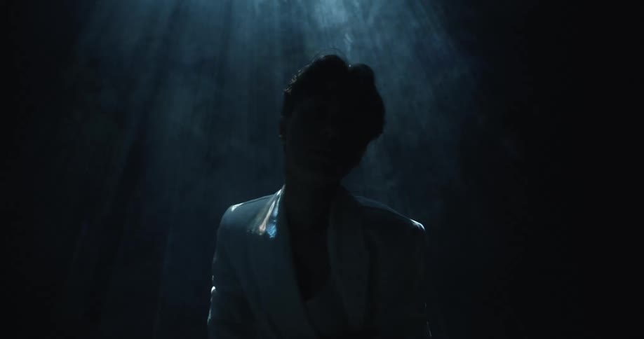 An asian woman wearing a white jacket and shirt stands in the dark with a strobing light and smoke in the air