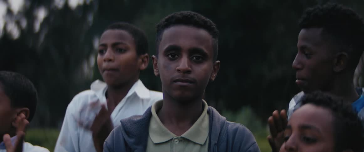 A teenage boy stands outside looking stoic at the camera while other boys jump and clap around him