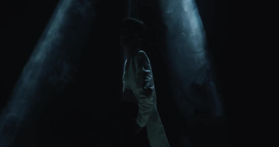 A thin asian female wearing a long white coat stands in the darkness under narrow spotlights
