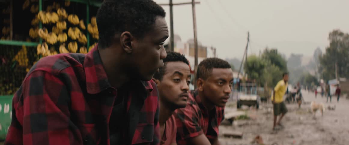 A young man comes to sit beside another pair of men beside a road in a city on a cloudy day