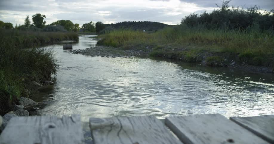 A rivers flows calmly under a simple wooden bridge on a partially cloudy day