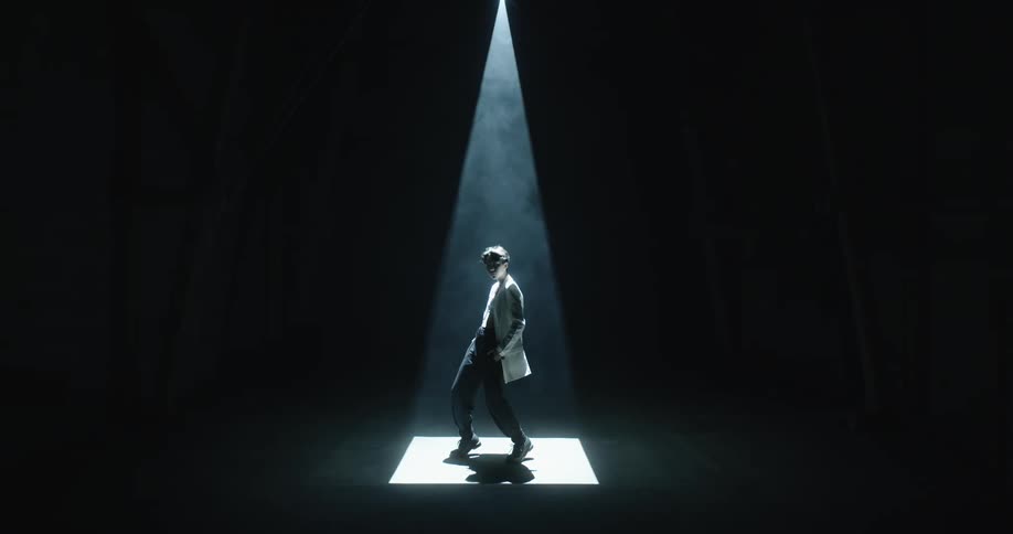Woman poses in widening spotlight beam in dark room with slanting wooden beams